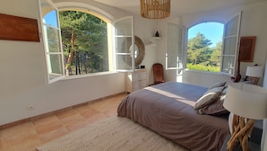 Un dormitorio con una cama grande, dos ventanas, una mesita de noche, una lámpara y un espejo.