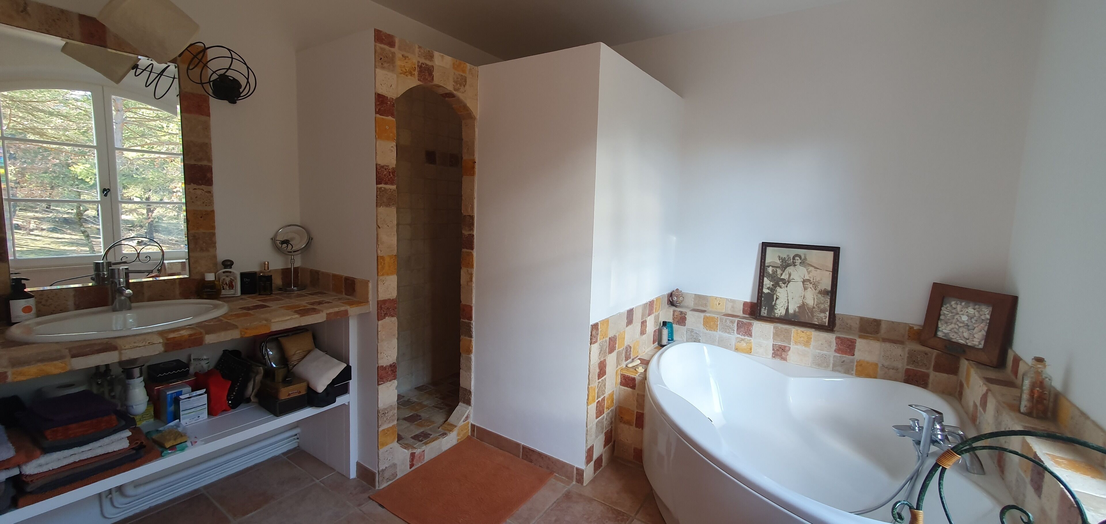 A bathroom with a large bathtub, a sink, and a mirror. There is a window providing natural light and a tiled wall with a unique pattern.