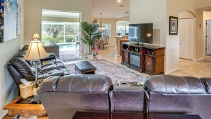A spacious living room with a leather sofa, a TV, and a dining area with a large window. - Beach Days & Fairways: Rotonda West Snowbird Home (Rotonda West)