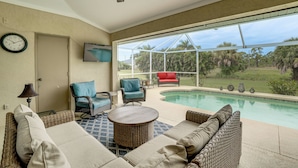 A poolside area with wicker furniture, a clock, and a TV on the wall. - Beach Days & Fairways: Rotonda West Snowbird Home (Rotonda West)