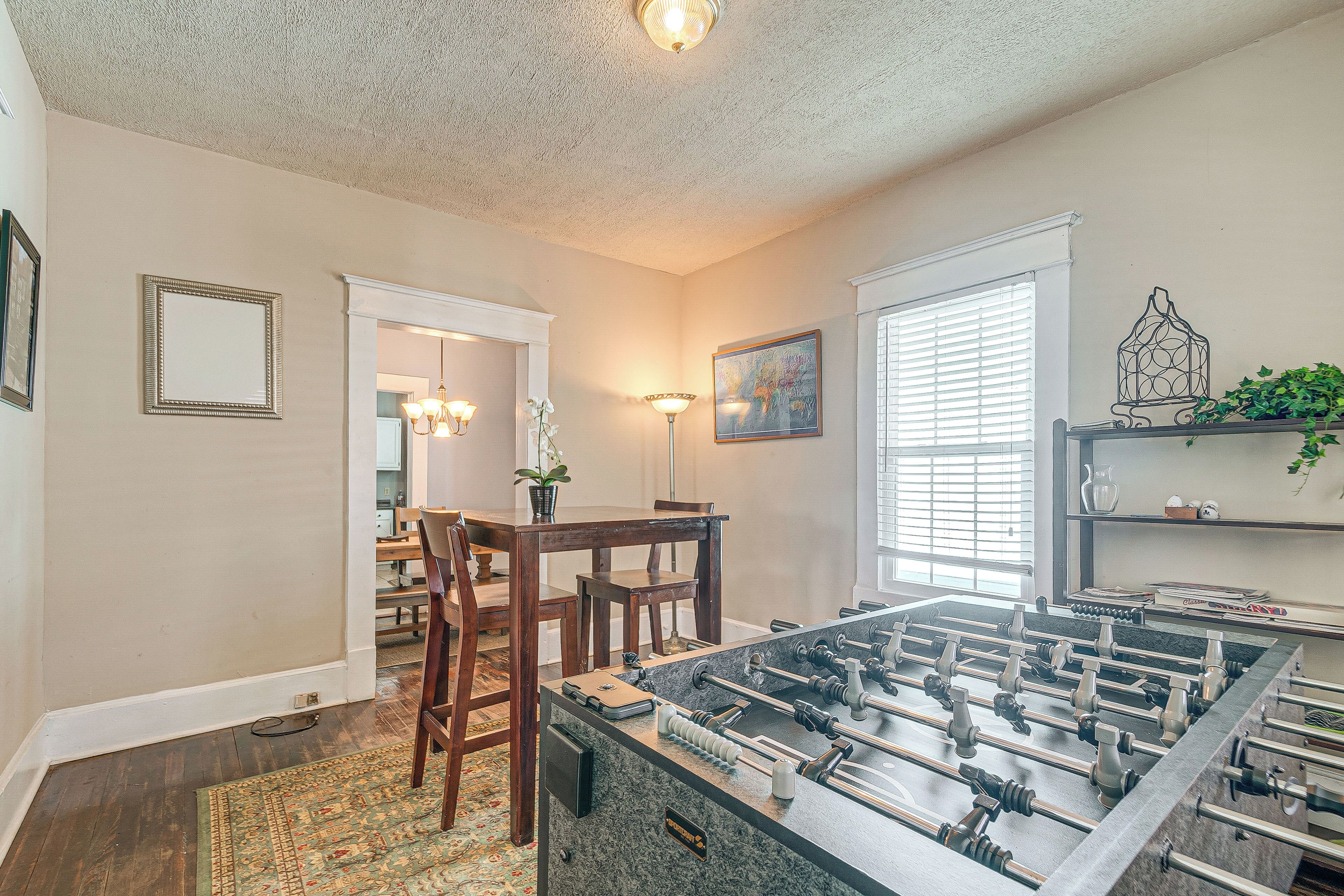 A room with a foosball table, wooden chairs, a dining table, and a chandelier.