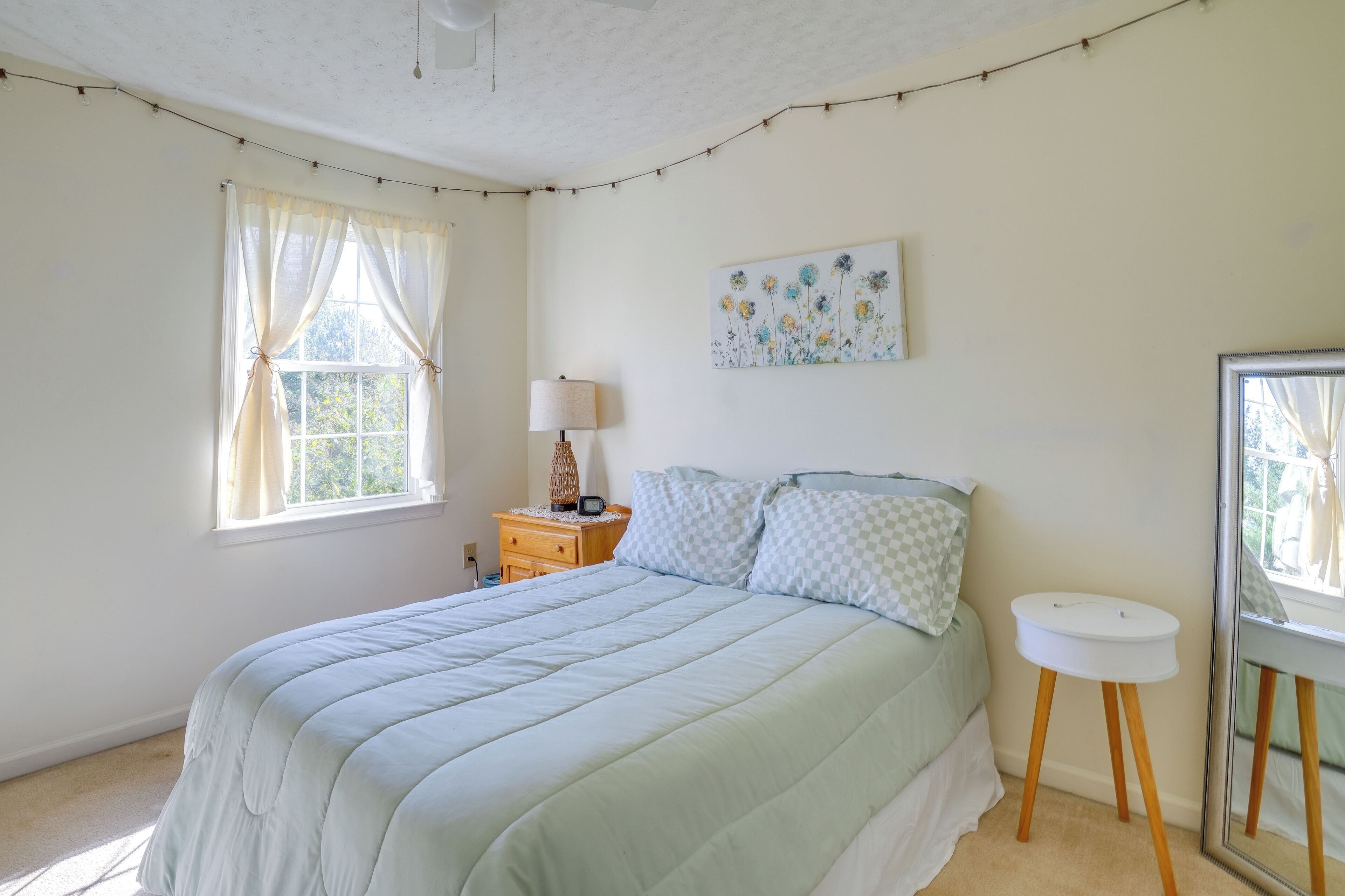 A bedroom with a bed, a bedside table, a lamp, a window with curtains, a wall-mounted painting, and a mirror.