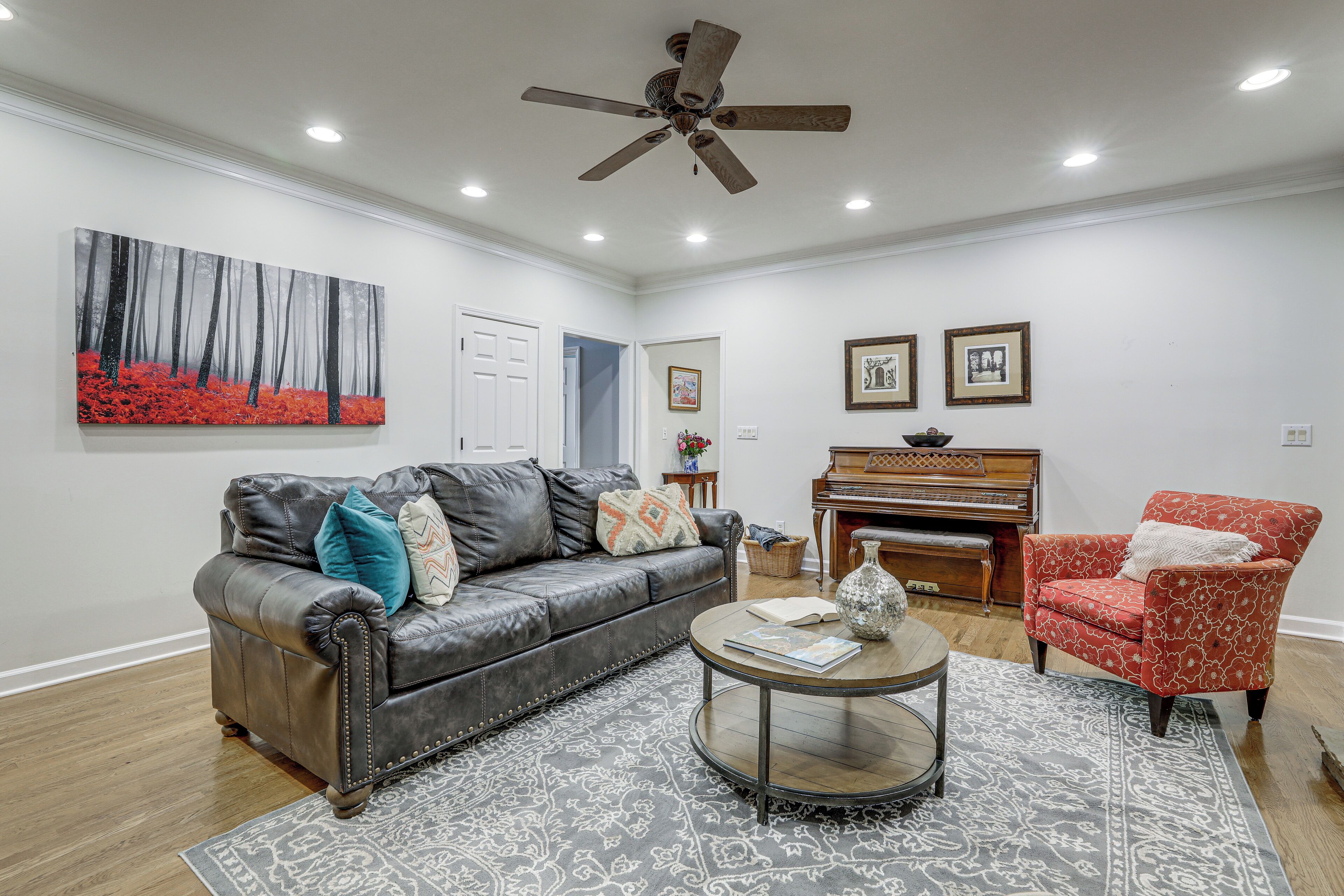 A living room with a leather sofa, a red armchair, a round coffee table, a piano, and a painting on the wall.