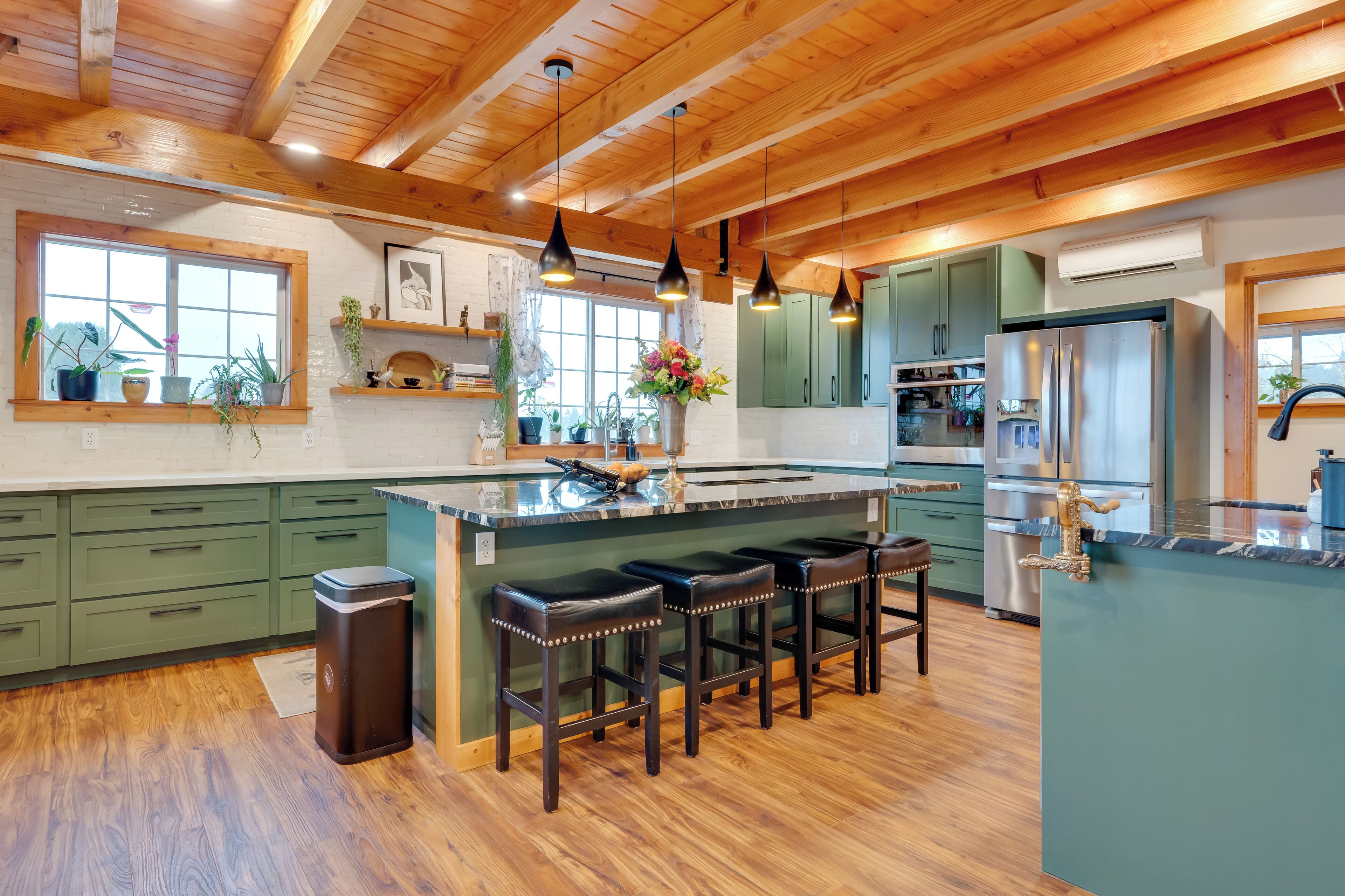 A spacious kitchen with wooden beams, green cabinets, a central island with stools, and a stainless steel refrigerator.