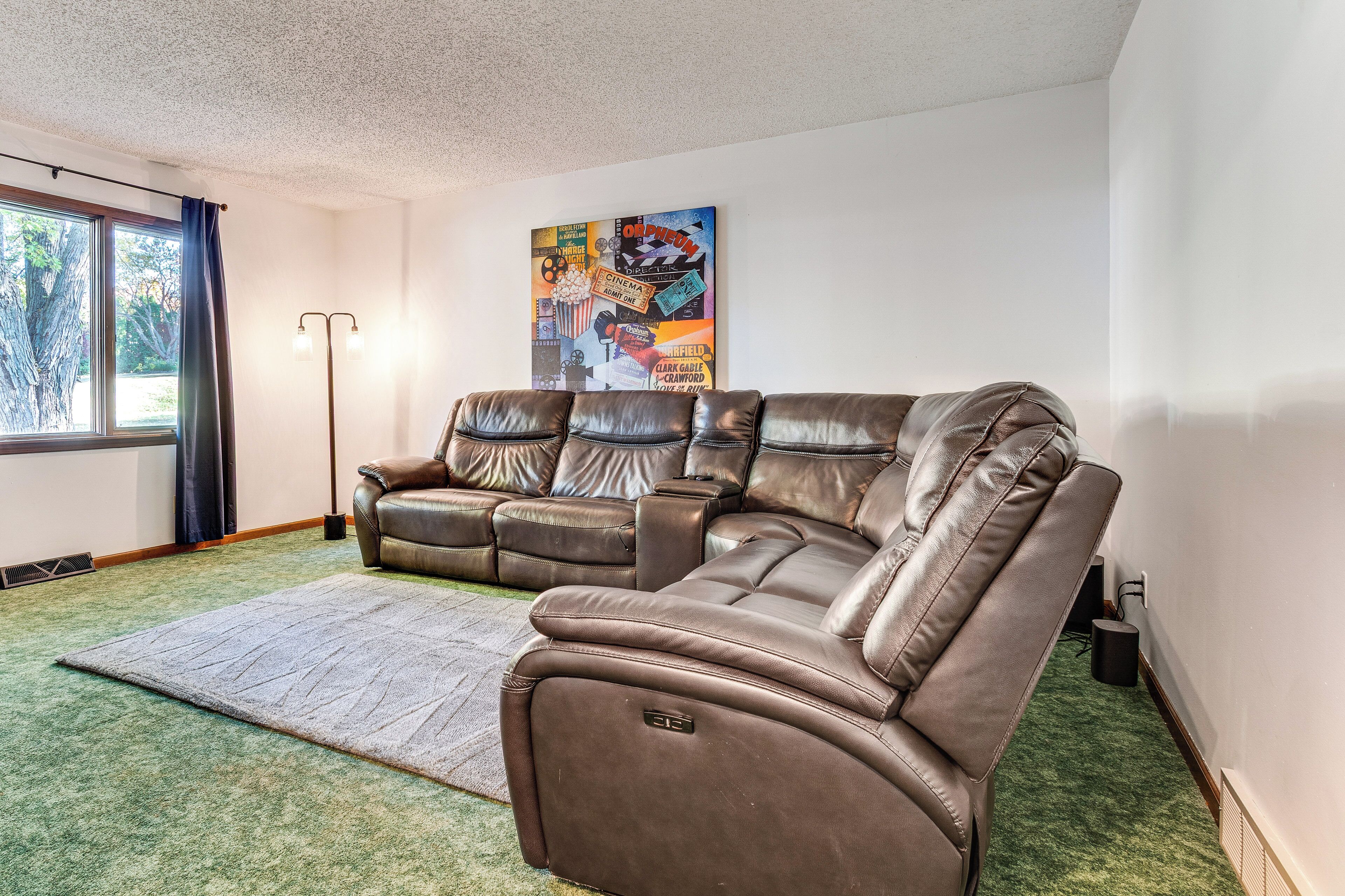 A living room with a brown leather sectional sofa, a green carpet, a large window with curtains, and a wall-mounted picture.