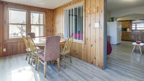 Een kamer met houten lambrisering, een eettafel en stoelen, een keuken met houten kasten, en een raam met zonwering.