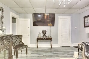 A room with a large flat-screen TV mounted on the wall, a wooden console table with decorative items, a tufted armchair, and a patterned sofa. - Canton Condo Near Football Hall of Fame & Dtwn! (Canton)