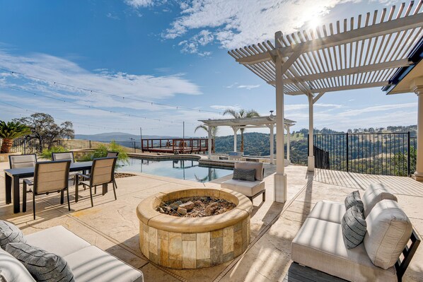 Zona junto a la piscina con un hogar, asientos exteriores y una pérgola con vistas a un paisaje pintoresco.