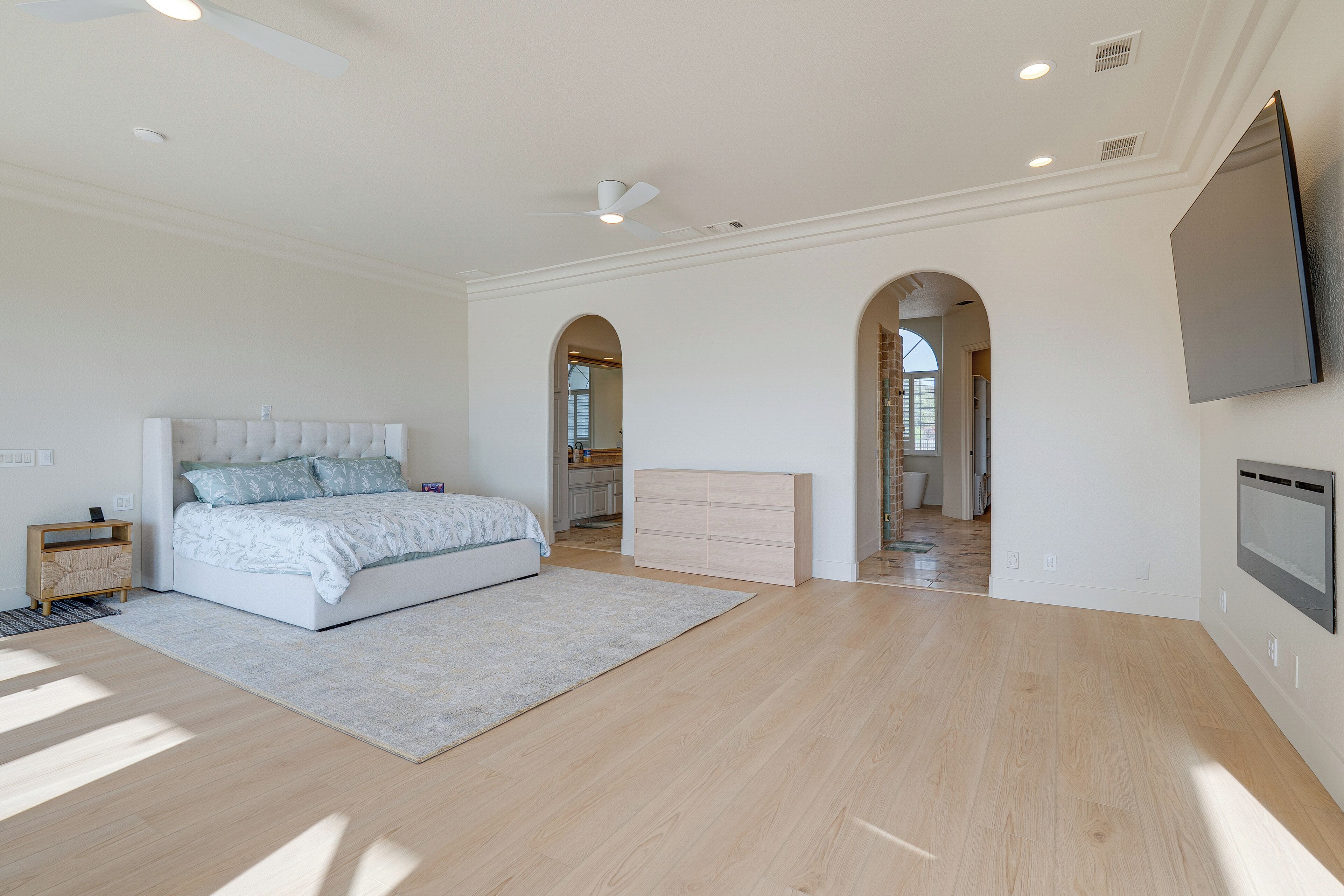 A spacious bedroom with a large bed, a wooden nightstand, a flat-screen TV mounted on the wall, and a ceiling fan.
