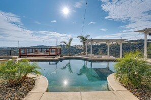 Una piscina de borde curvo, rodeada de rocas y plantas de palma. Hay una terraza de madera con sillas y una pérgola.