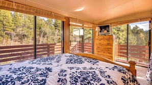 A bedroom with a large bed, wooden dresser, and a view of trees through large windows. - Close to Pike Natl Forest! Lake-view Family Cabin (Divide)