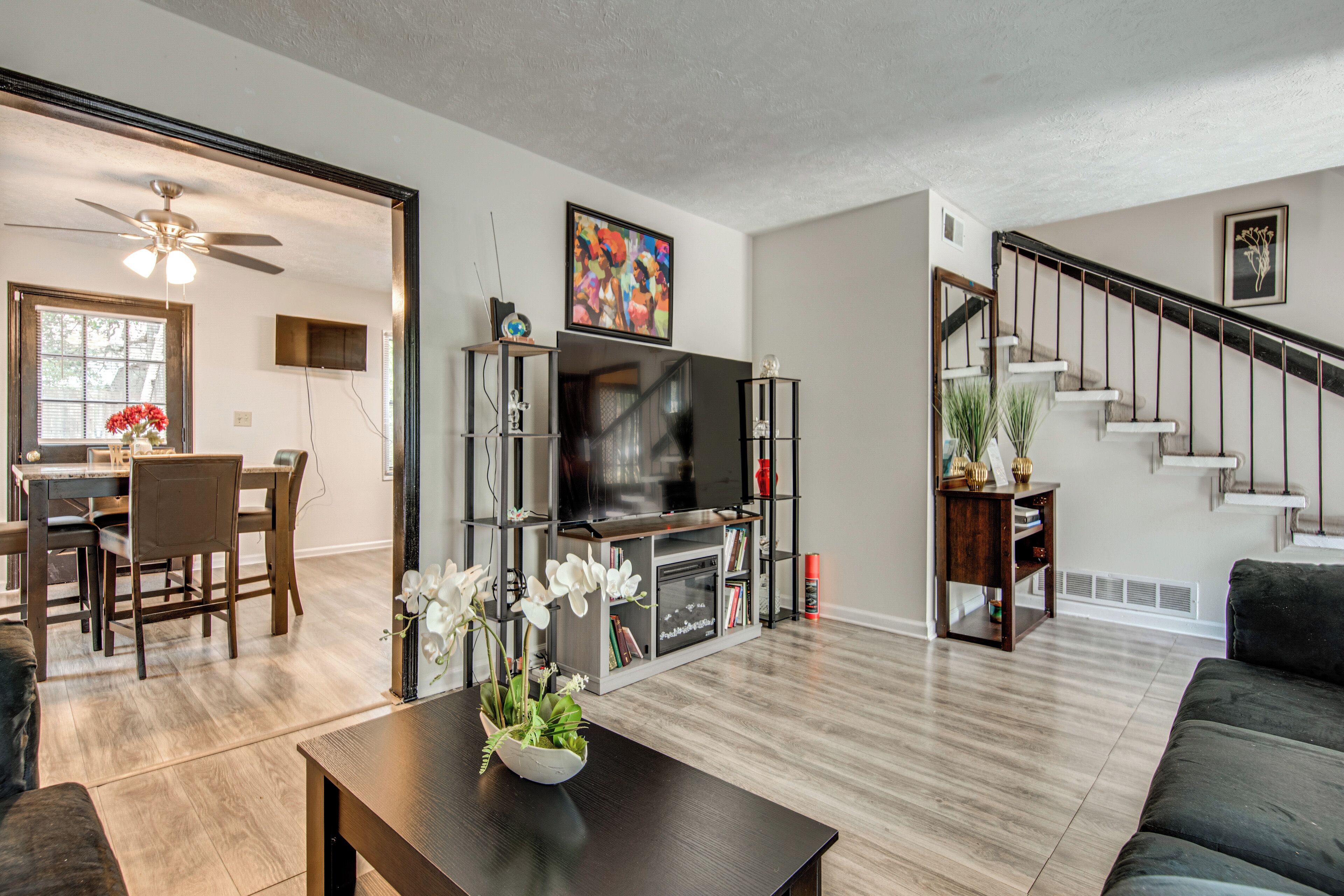 A modern living room with a flat-screen TV, a dining area with a wooden table and chairs, and a staircase leading to another level.