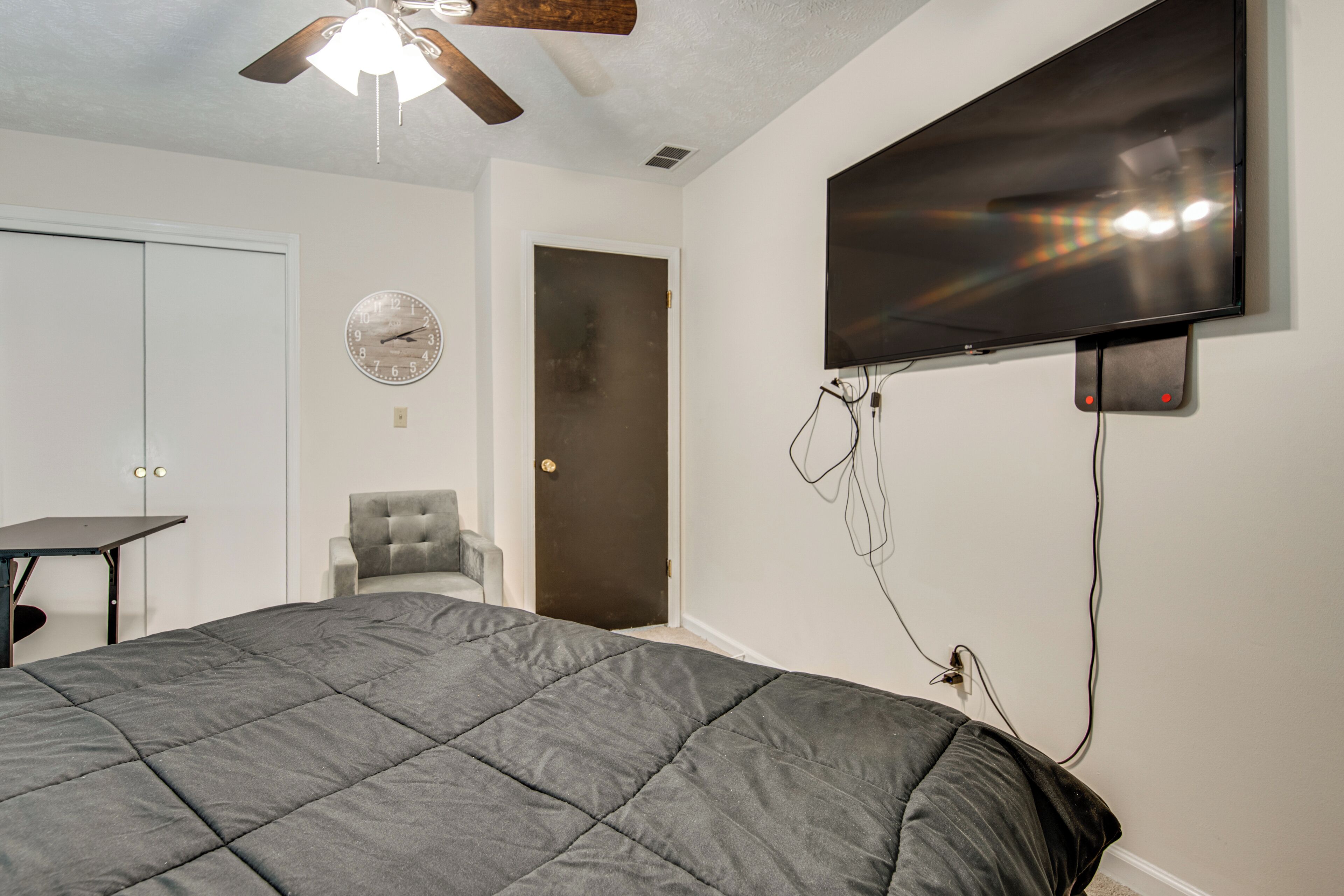 A bedroom with a bed, a chair, a desk, a TV mounted on the wall, and a ceiling fan.