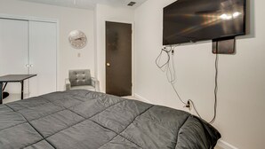Quarto com cama, poltrona, escrivaninha, televisão fixada na parede e ventoinha no teto.