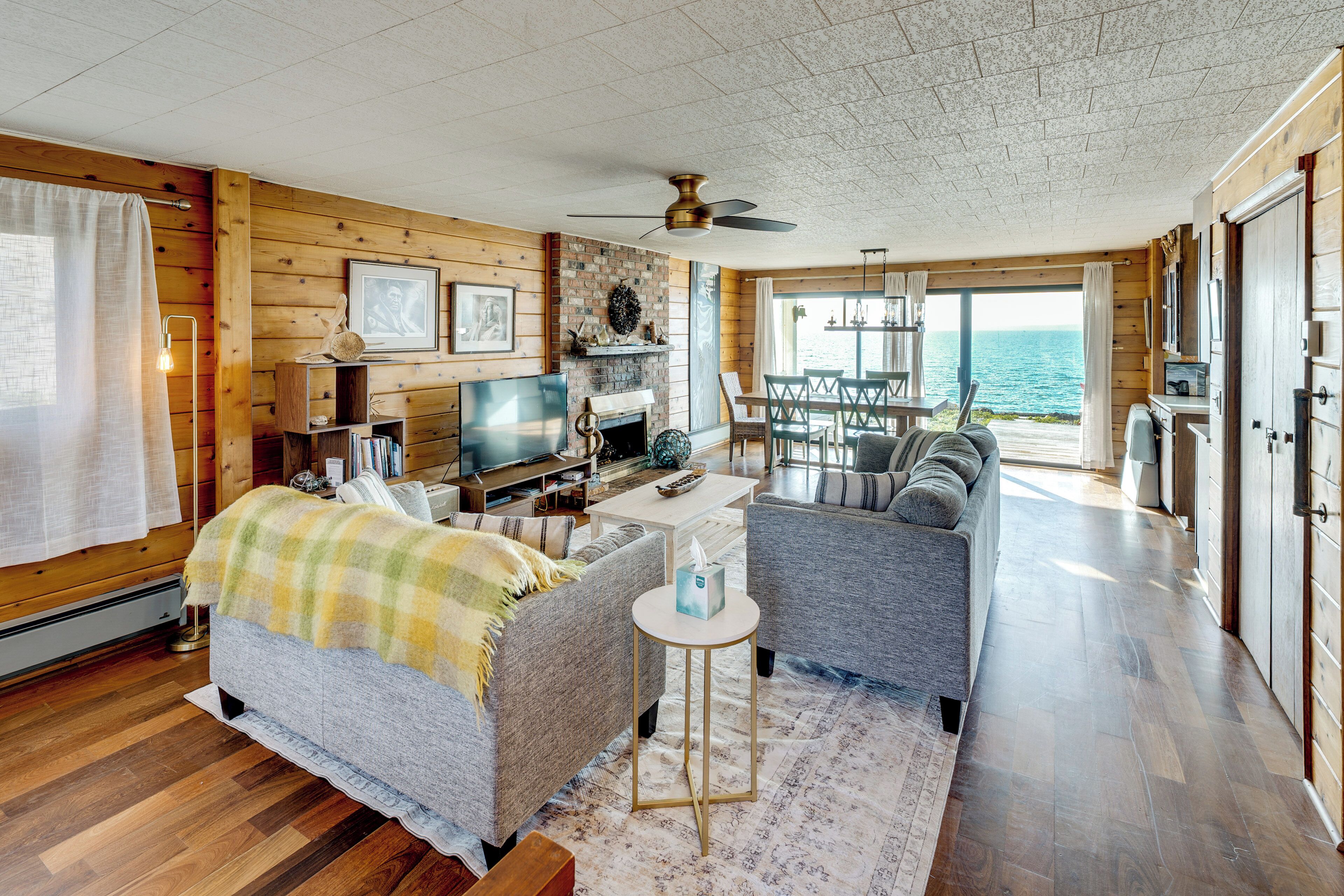 A cozy cabin interior with a fireplace, wooden walls, a dining area, and a view of the ocean.