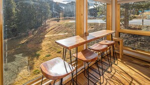 A wooden bar area with a view of a forested hillside and a building in the distance. - Beautiful Mountain Views, walking distance to Big Sky Resort! (Big Sky)