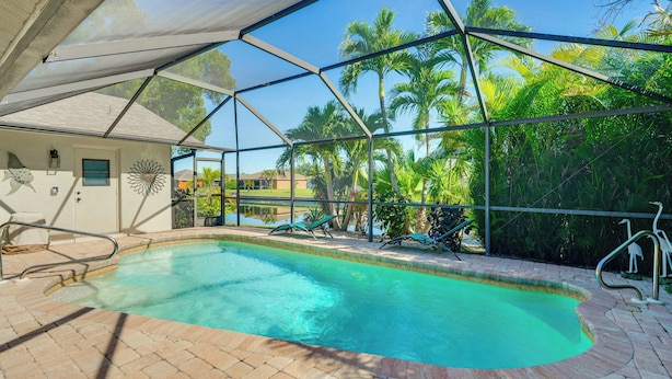 A swimming pool with a clear blue water, surrounded by a covered area with a view of palm trees and a house in the background. - Canal-front Escape w/ Dock & Pool in Cape Coral (Cape Coral)