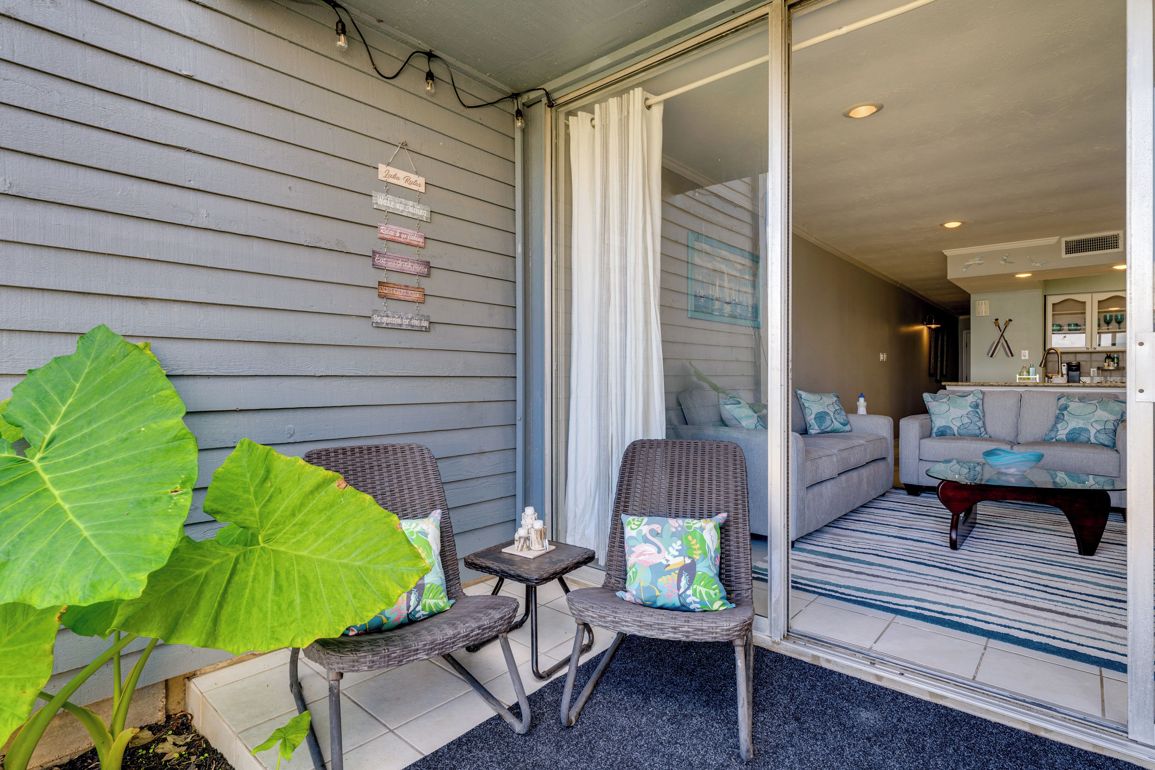 A covered outdoor seating area with wicker chairs and a small table, a sliding glass door leading to a living room with a sofa and a coffee table.