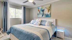 Un dormitorio con cama, ventilador de techo, mesita de noche y ventana con cortinas.