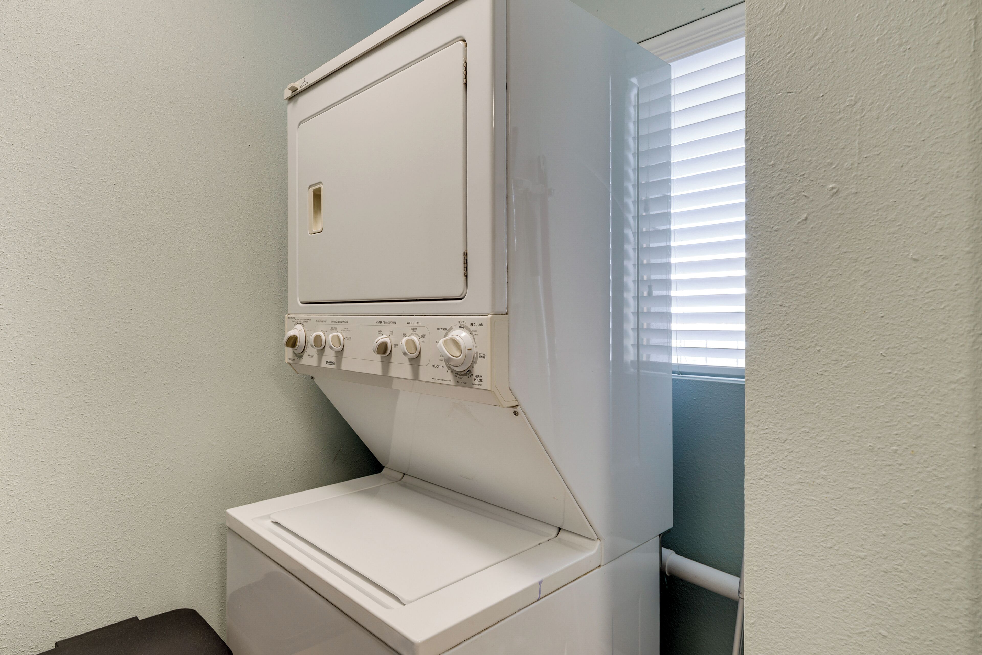 A compact laundry room with a washing machine and dryer, a small trash bin, and a window with blinds.