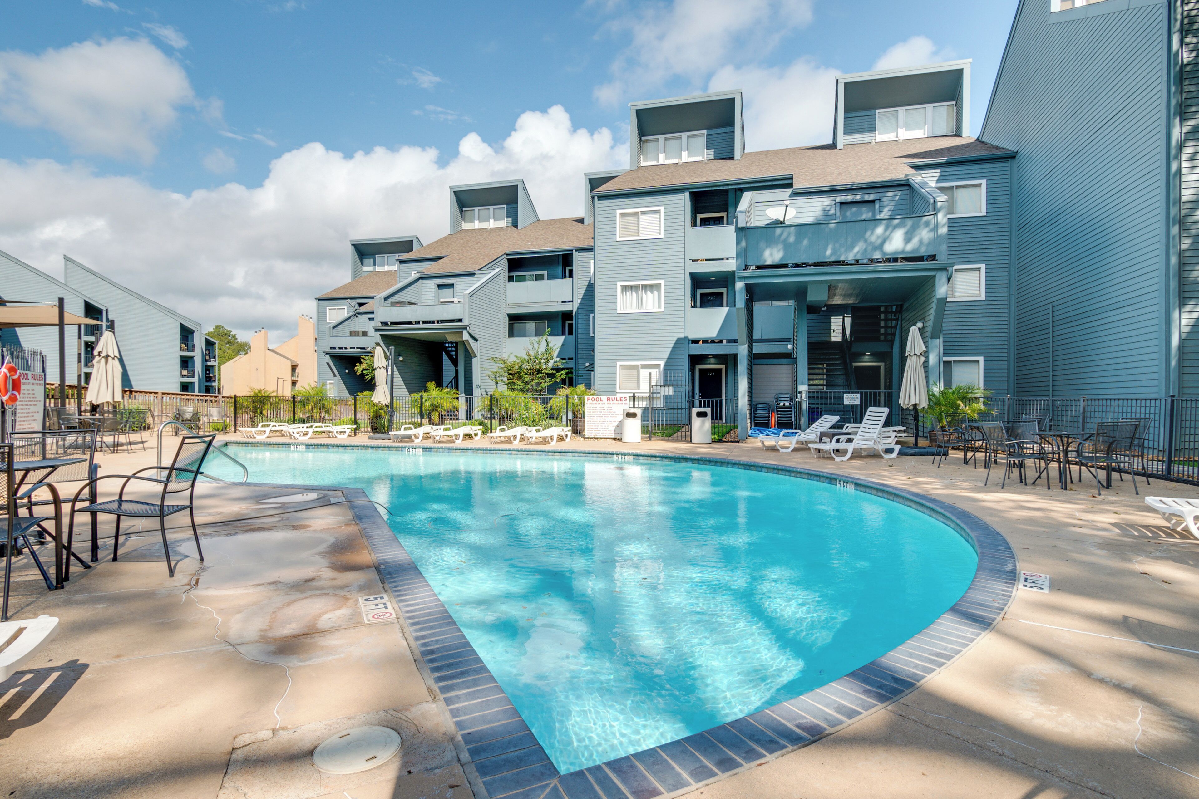 A swimming pool surrounded by modern buildings with balconies, lounge chairs, and a clear blue sky.
