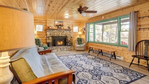 A cozy cabin interior with a stone fireplace, wooden walls, a patterned rug, and a large window with curtains. - Hike On-site! 16-acre Escape Near Shenandoah NP (Stanley)