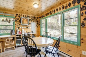 A wooden cabin interior with a dining table, chairs, a kitchen area, and a large window offering a view of the outdoors.