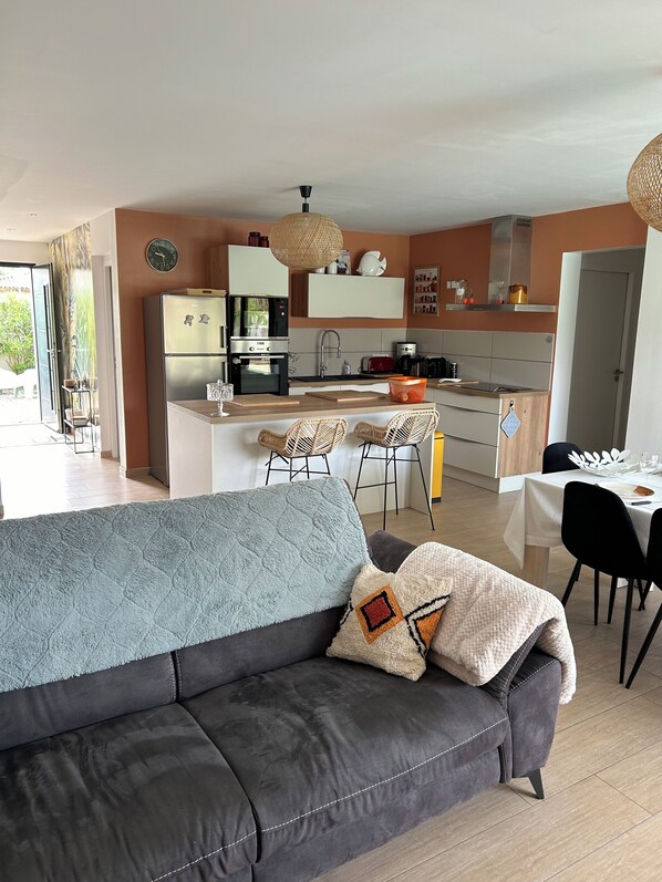 Ein modernes Wohnzimmer mit grauem Sofa, einem Essbereich mit Tisch und Stühlen sowie einer Küche mit Geräten und einer Pendelleuchte.