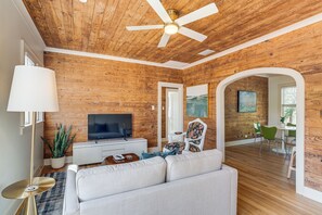 A modern living room with a wooden ceiling, a white sofa, a television, and a dining area with green chairs. - Walk to Downtown Tyler! Updated Historic Apt (Tyler)