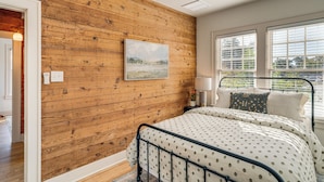 A bedroom with a wooden wall, a bed with a patterned bedspread, a window with blinds, and a painting on the wall. - Walk to Downtown Tyler! Updated Historic Apt (Tyler)