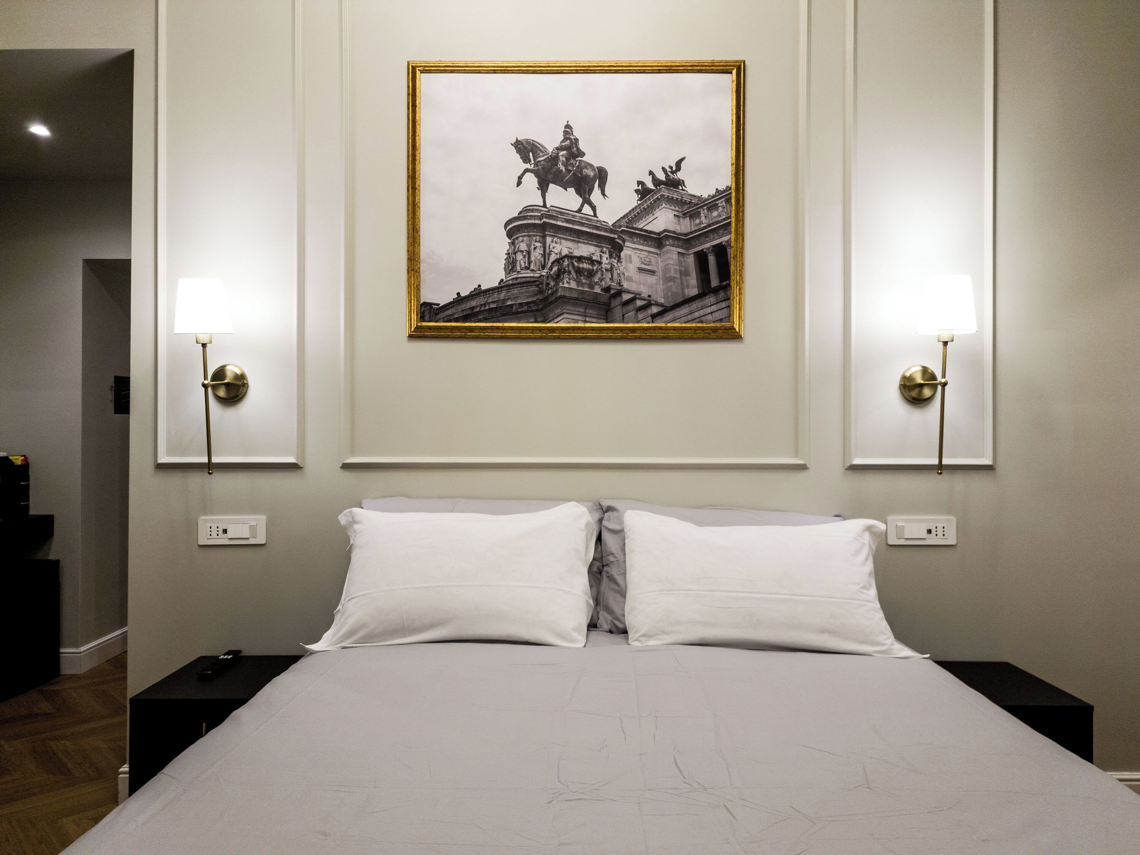 Une chambre d’hôtel avec un grand lit, deux tables de chevet, une photographie en noir et blanc encadrée accrochée au mur, et deux lampes fixées au mur.