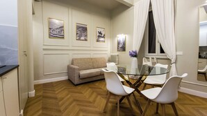 A dining area with a glass table, white chairs, a beige sofa, and three framed pictures on the wall. - Domus Innovatis Rooms & Home (Rome)