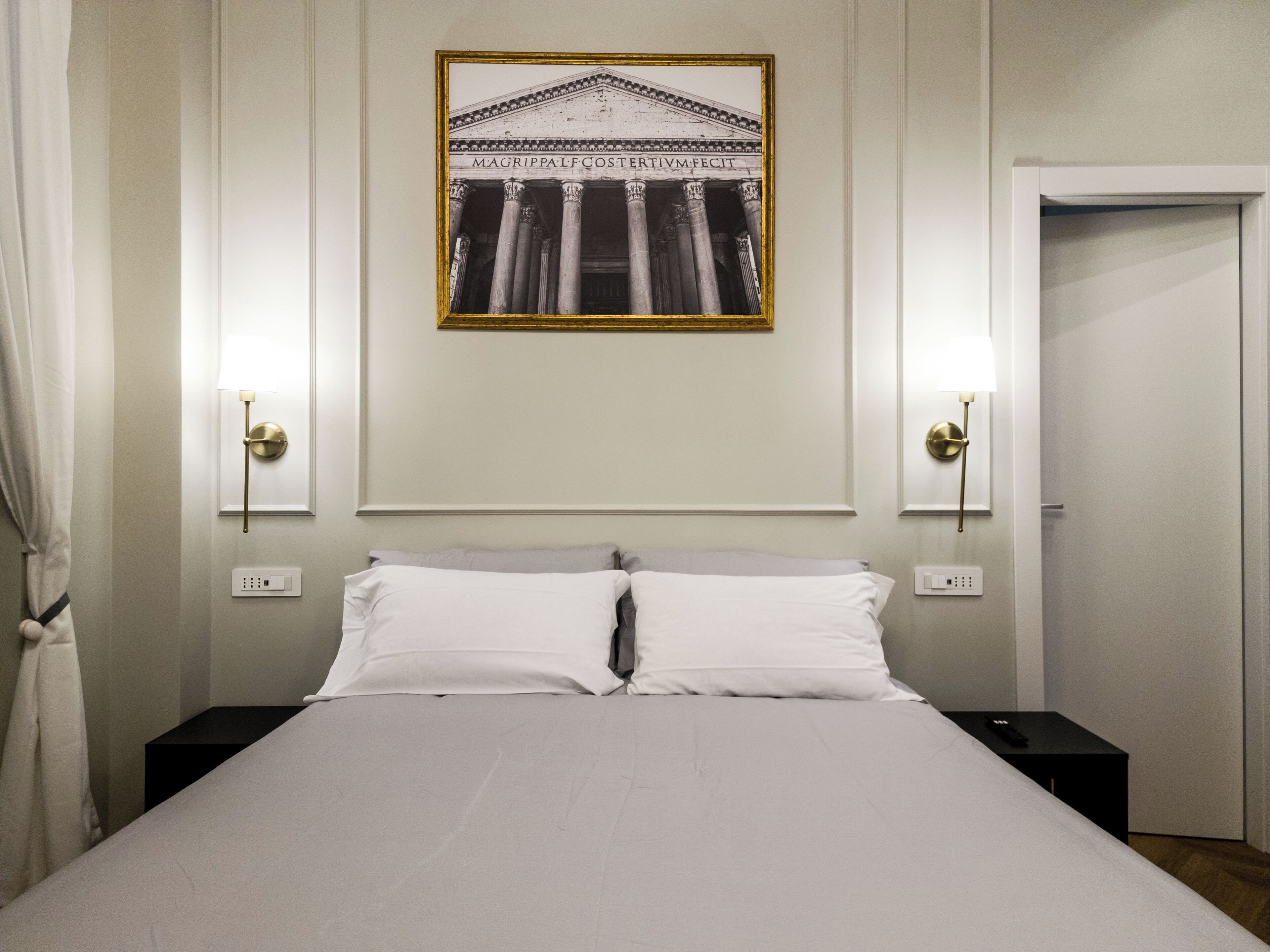 En välordnad säng med vita sängkläder i ett hotellrum. På väggen ovanför sängen hänger ett inramat svartvitt foto av en klassisk byggnad.