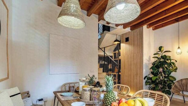 Un comedor con una mesa de madera dispuesta para cuatro personas, rodeada de sillas de mimbre. Hay una cesta de frutas en la mesa y una planta en maceta en la esquina.