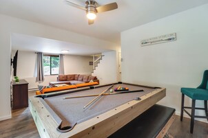 Una mesa de billar en una habitación con ventilador de techo, un sofá y una escalera que sube a otro nivel.