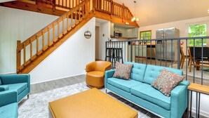 A spacious living area with a wooden staircase, teal sofa, orange armchair, and a kitchen in the background. - 1 Mi to Lake, Golf & Ski Access: The Hideout Home! (Lake Ariel)