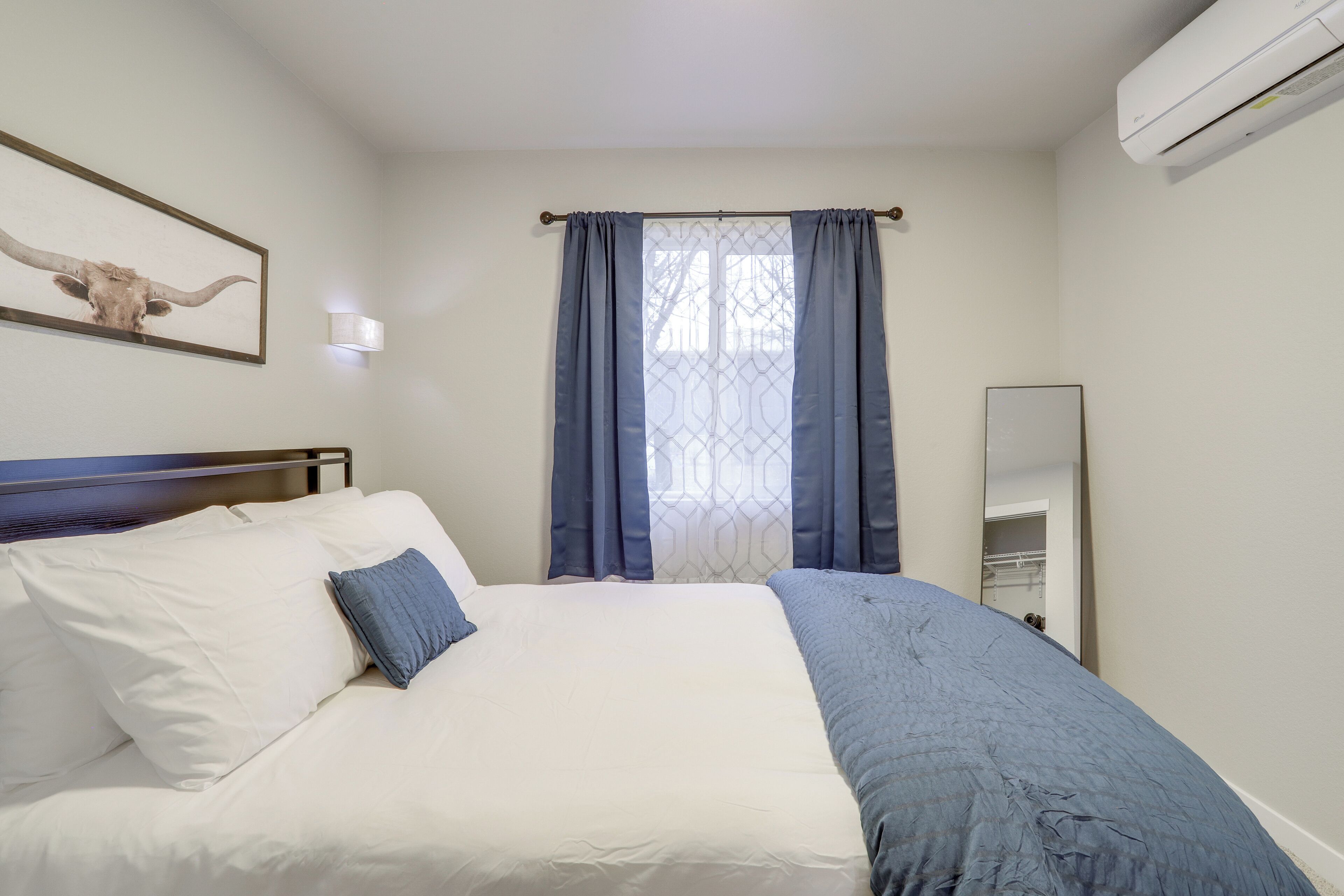 A neatly made bed with white and blue bedding, a wall-mounted light, a framed picture, and a window with blue curtains.