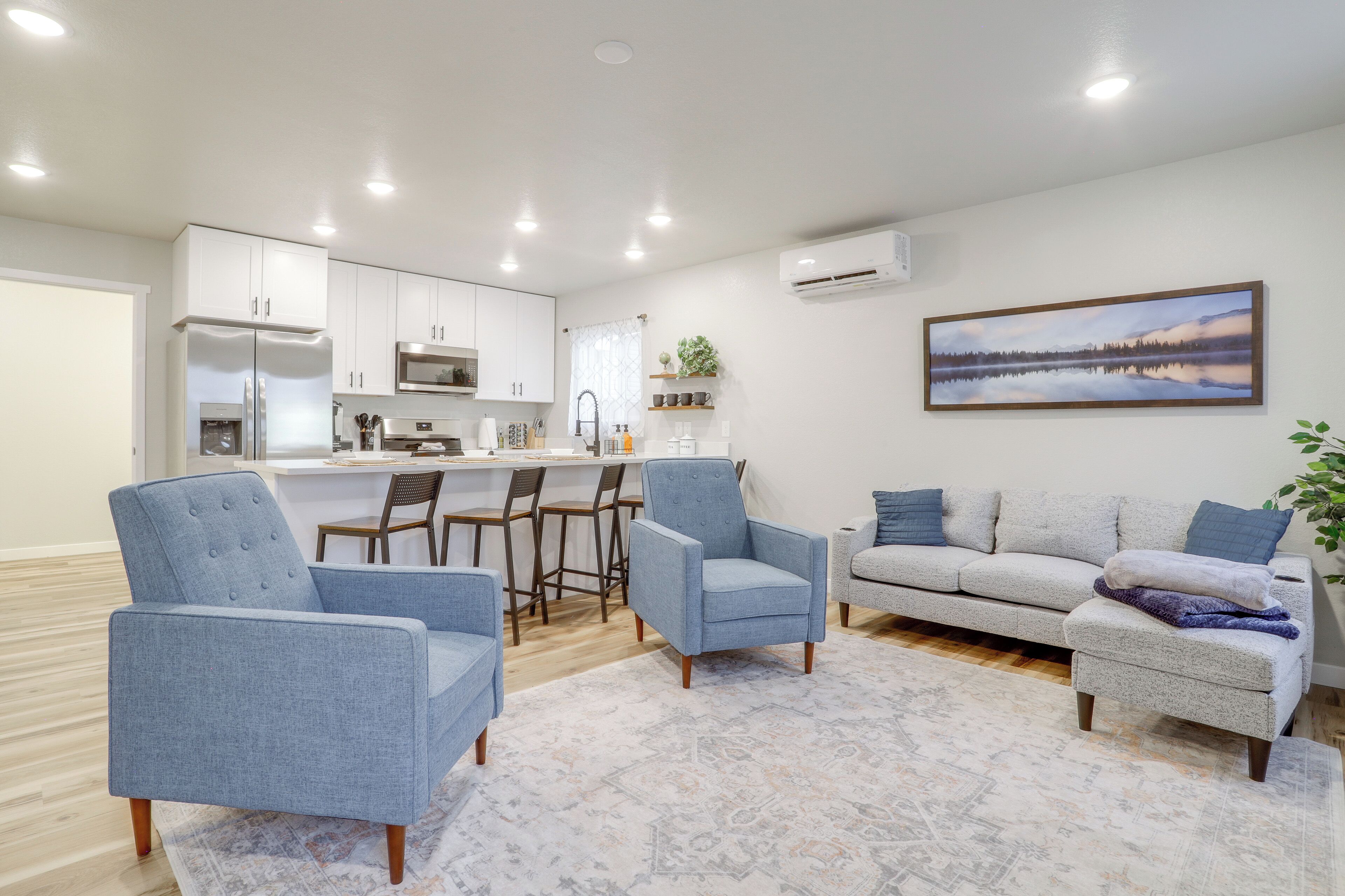 A modern living room with a sofa, two armchairs, and a coffee table. There is a kitchen area with white cabinets and a refrigerator. A painting of a lake is on the wall.