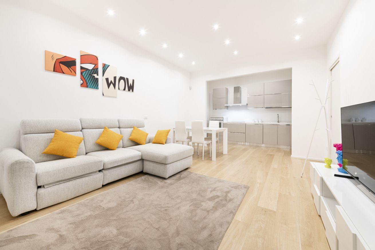 A modern living room with a grey sectional sofa, a flat-screen TV, and a kitchen in the background.