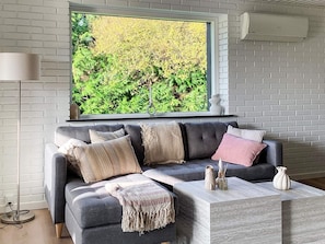 Ein modernes Wohnzimmer mit grauem Sofa, einem großen Fenster mit Blick auf üppiges Grün, einer weißen Ziegelwand und einem Marmortisch.