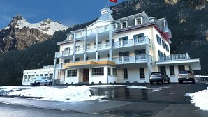 A multi-story hotel with balconies, a red flag, and a mountainous backdrop. - Gemmi Hotel & Spa (Kandersteg)