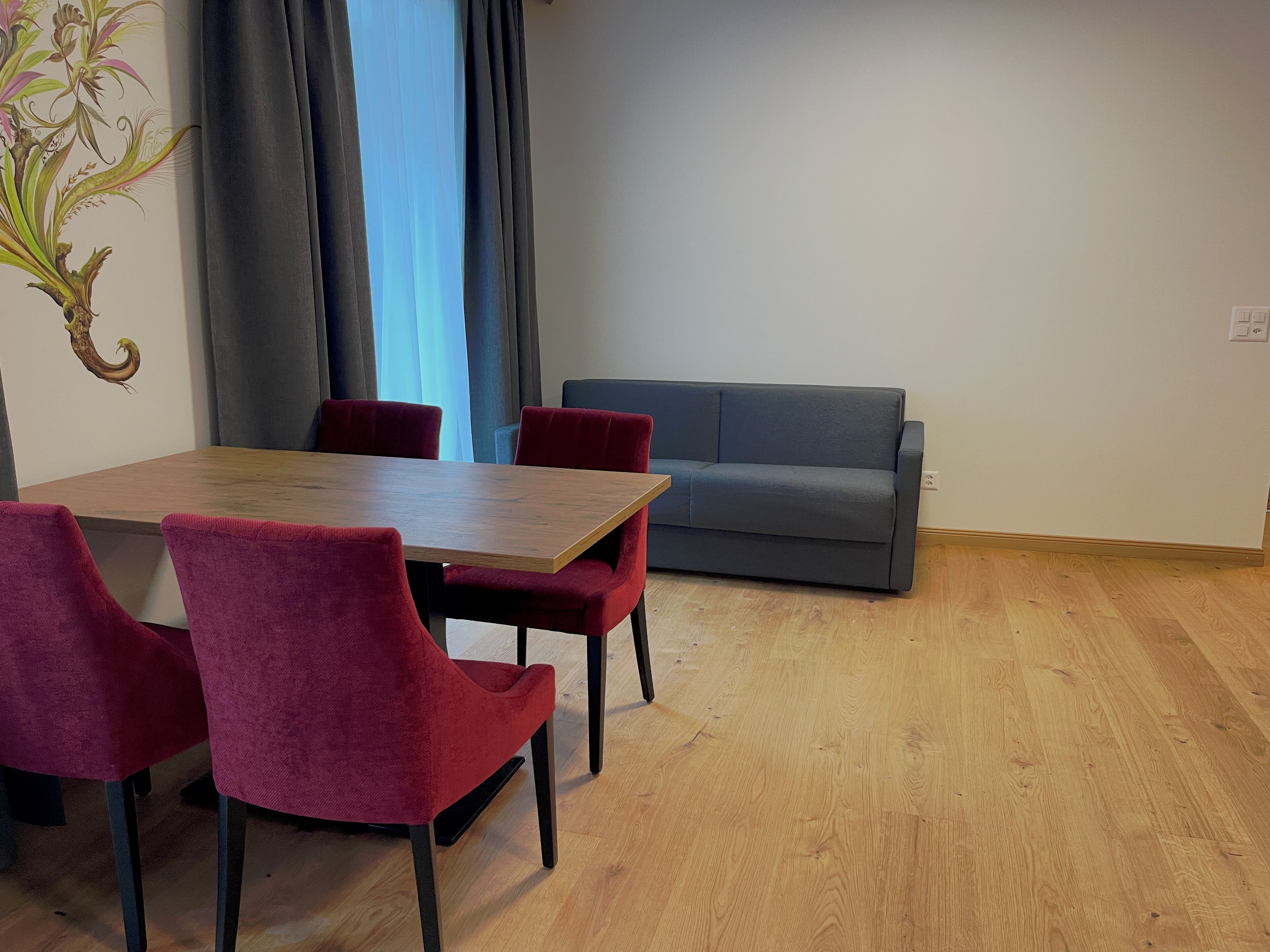 Un comedor con una mesa de madera y sillas rojas, un sofá gris y un ventanal con cortinas.