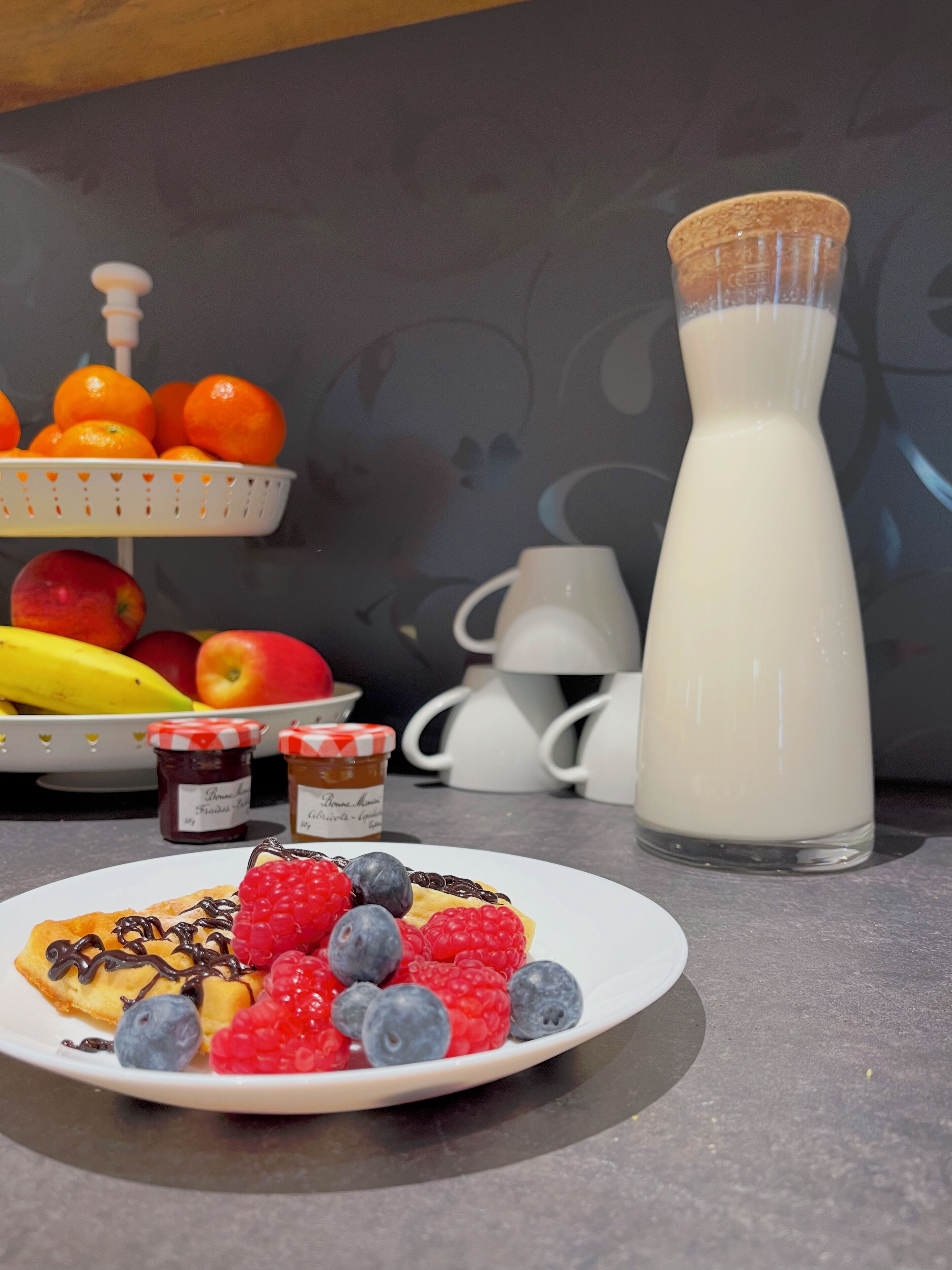 A breakfast setting with a plate of pancakes topped with berries and chocolate sauce, a milk bottle, and a fruit basket with oranges and apples.