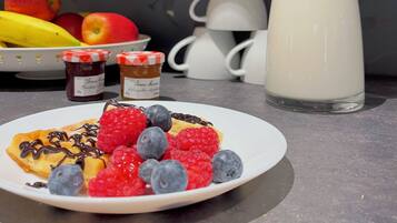 A breakfast setting with a plate of pancakes topped with berries and chocolate sauce, a milk bottle, and a fruit basket with oranges and apples.