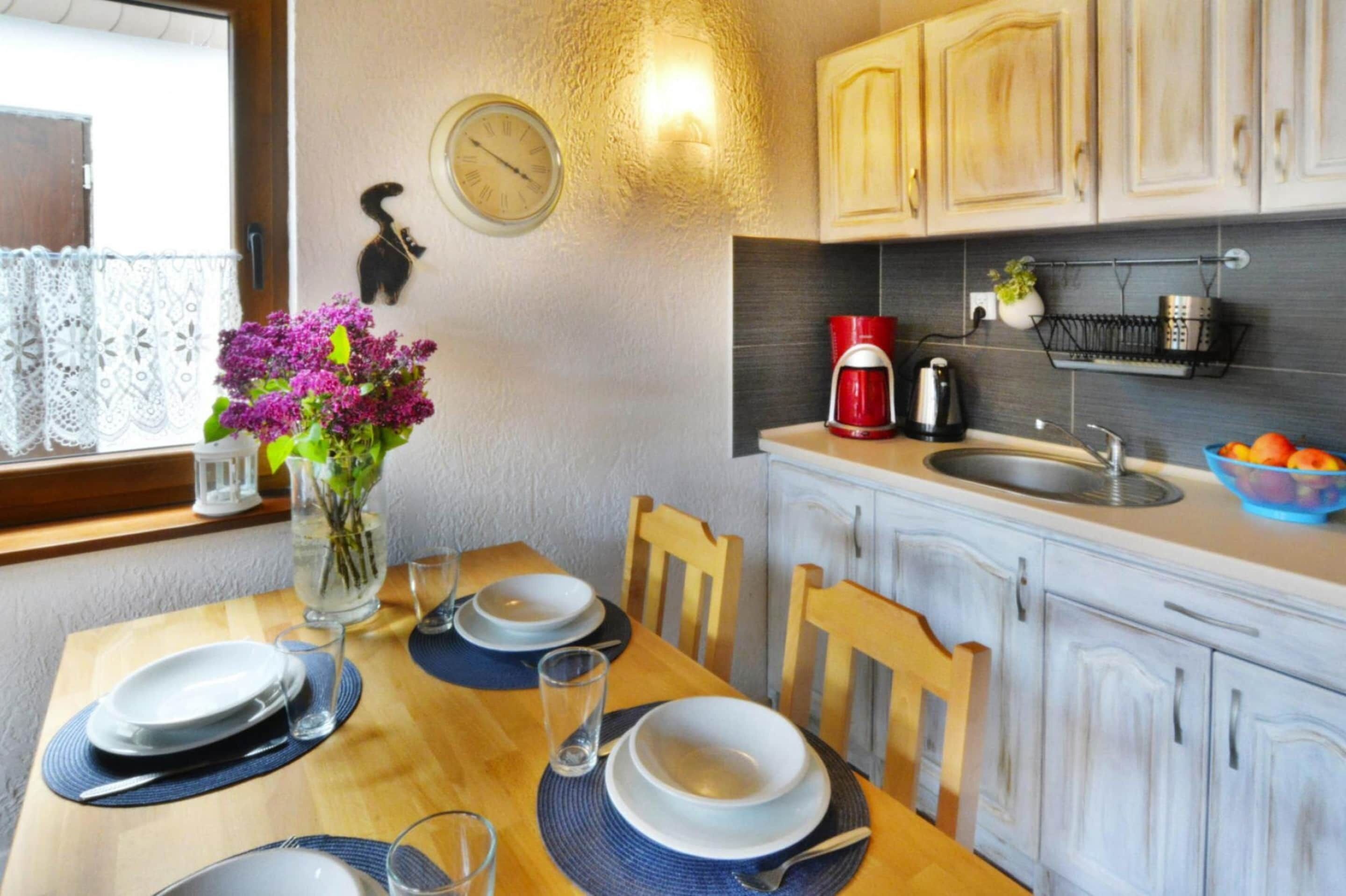 Un coin repas avec une table en bois dressée pour deux personnes, un vase rempli de fleurs violettes, une horloge au mur et des meubles de couleur claire.