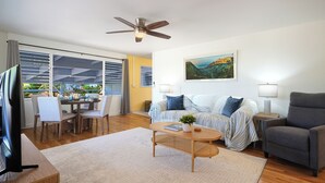 A living room with a ceiling fan, a dining area with a table and chairs, a sofa with a plant, and a painting on the wall. - Charming 3BR Koloa Shore Cottage with ocean views and AC, sleeps 6 in Poipu (Koloa)
