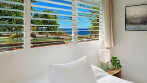 A bedroom with a bed, a nightstand with a lamp, a potted plant, and a view of the ocean through window blinds. - Charming 3BR Koloa Shore Cottage with ocean views and AC, sleeps 6 in Poipu (Koloa)