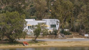 Una casa moderna con fachada blanca, contraventanas verdes y una escalera de piedra que lleva a un porche. Un barco está amarrado en la orilla, y se ve un letrero que dice "Casa de Andes".