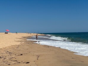 Une plage avec un ciel dégagé, des gens qui se promènent le long du rivage et des parasols installés pour se protéger du soleil.
