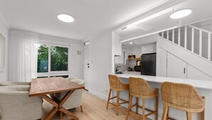 A modern kitchen with white cabinetry, a wooden dining table, and wicker bar stools. - Stylish 2-bed Townhouse in Leafy Hyde Park (Adelaide)